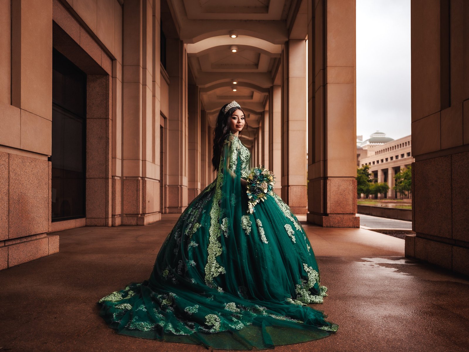 Quinceanera in regal emerald gown with long train, cinematic fine art portrait under grand columns.