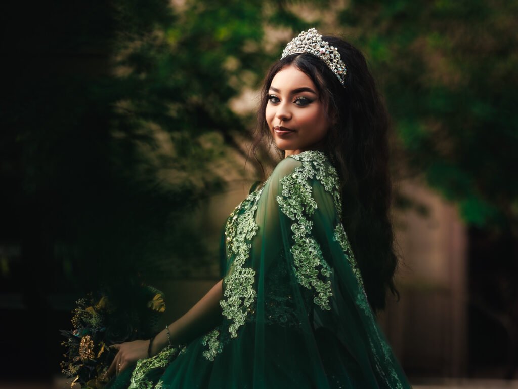 Quinceanera in emerald gown with lace cape looking back, dramatic moody lighting from a rainy day shoot near the Indiana State Capitol.