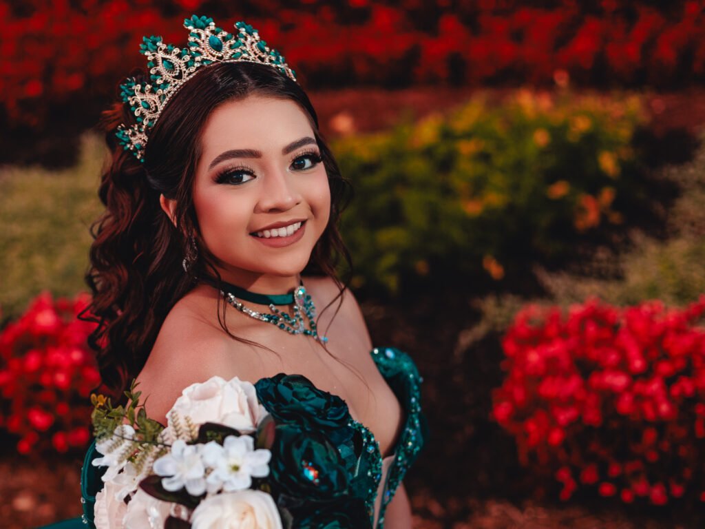Close-up portrait of smiling Quinceanera with tiara, soft cinematic lighting and red floral bokeh background at Coxhall Gardens.