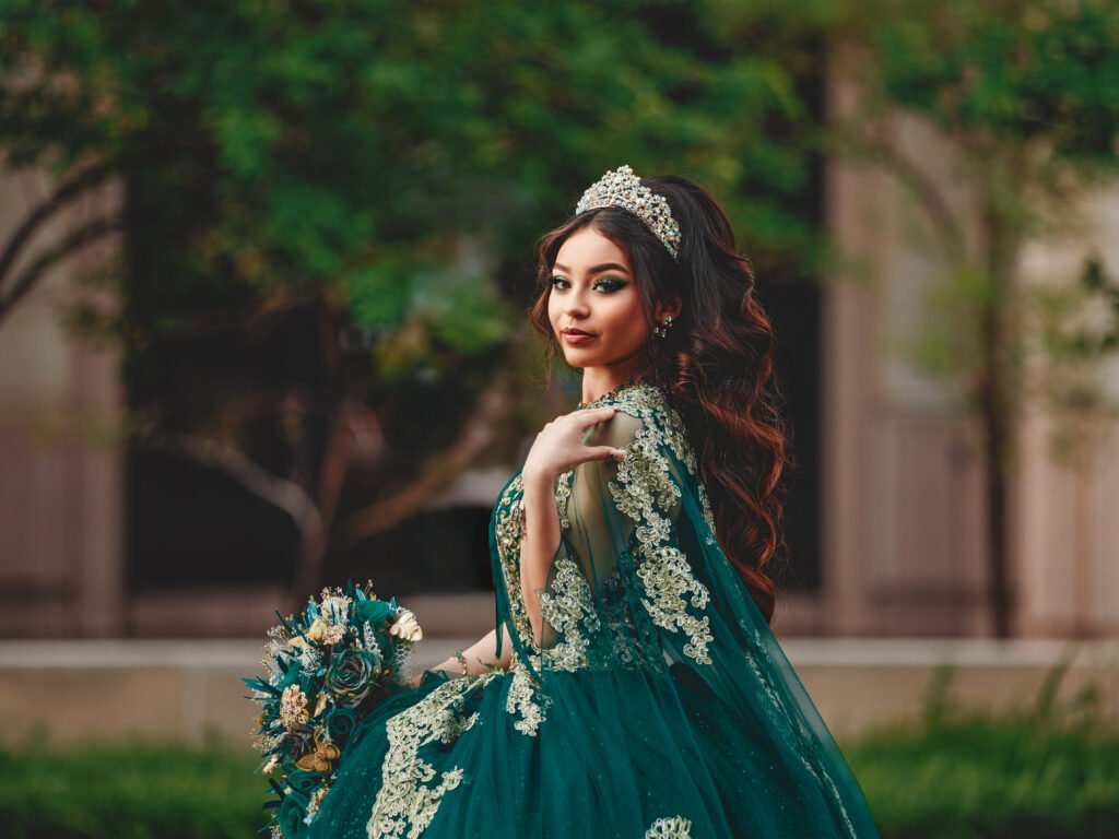Stunning portrait of a Quinceañera in an Emerald Green Quinceañera Dress with a long train and gold embroidery, standing with the Indiana State House in the background.