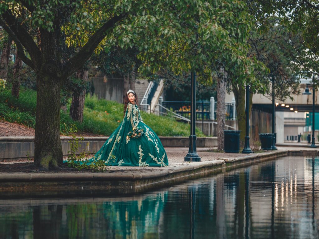 Beautiful Quinceañera in her Emerald Green Quinceañera Dress posing by the water at the Indianapolis Canal Walk, with trees and a serene reflection.