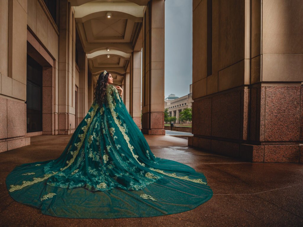 Captivating wide-angle shot of a Quinceañera in an Emerald Green Quinceañera Dress with a long train, featuring the Indiana State House in the background.