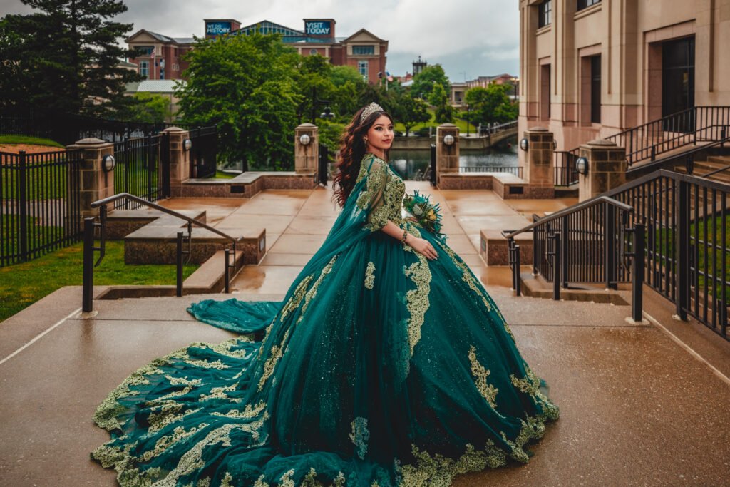 High-angle shot of a Quinceañera in an Emerald Green Quinceañera Dress standing at the top of a stone staircase that descends to the Indianapolis Canal Walk, with a bridge, buildings, and lush green trees in the background.