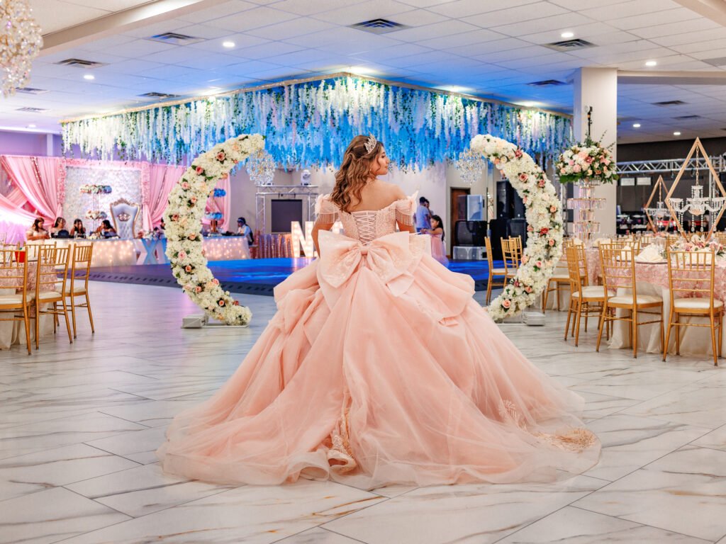 Quinceanera in blush gown, dramatic back bow, cinematic lighting, and elegant floral arches.