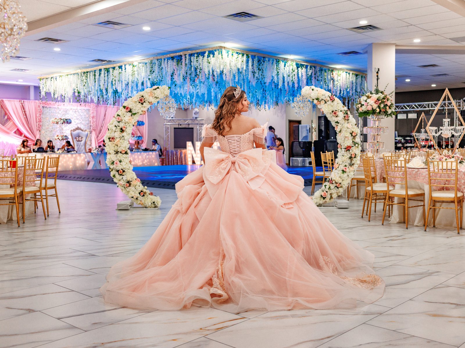Quinceanera in blush gown, dramatic back bow, cinematic lighting, and elegant floral arches.