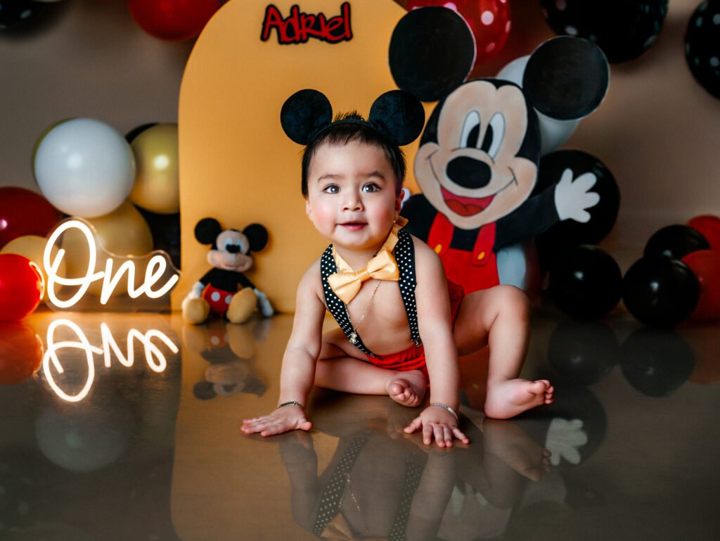 Happy smiling one-year-old boy sitting in a Mickey Mouse themed studio set with a custom "Adriel" backdrop and cake