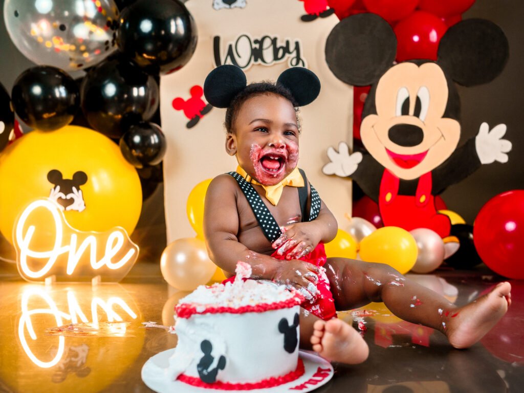 Niño de un año vestido de rojo sentado en un set de Mickey Mouse con globos y letrero de neón, fotografía de estudio profesional en Indianápolis.