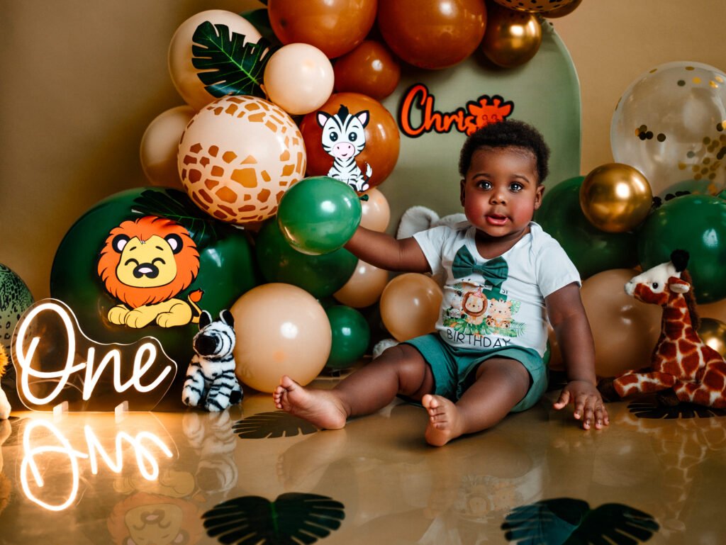 Toddler sitting in a jungle-themed safari set with a lion plush, giraffe toy, and an organic green and gold balloon arch.