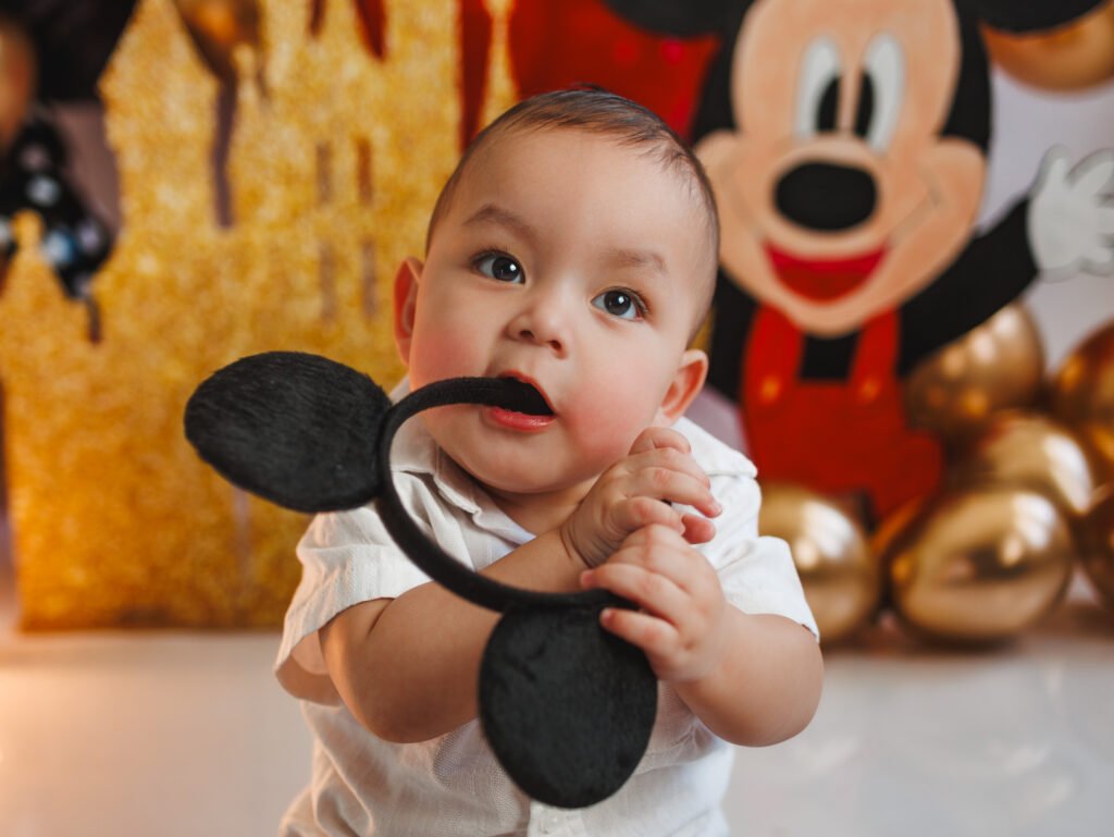 Cute one-year-old boy playing with Mickey ears during his Mickey Mouse Cake Smash Photography session.