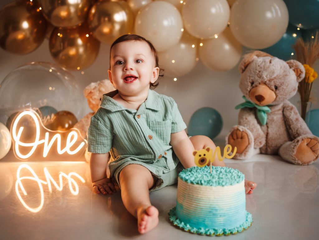 Smiling baby boy with blue smash cake, neon 'One' sign and teddy bear, cinematic warm lighting in Indianapolis studio.